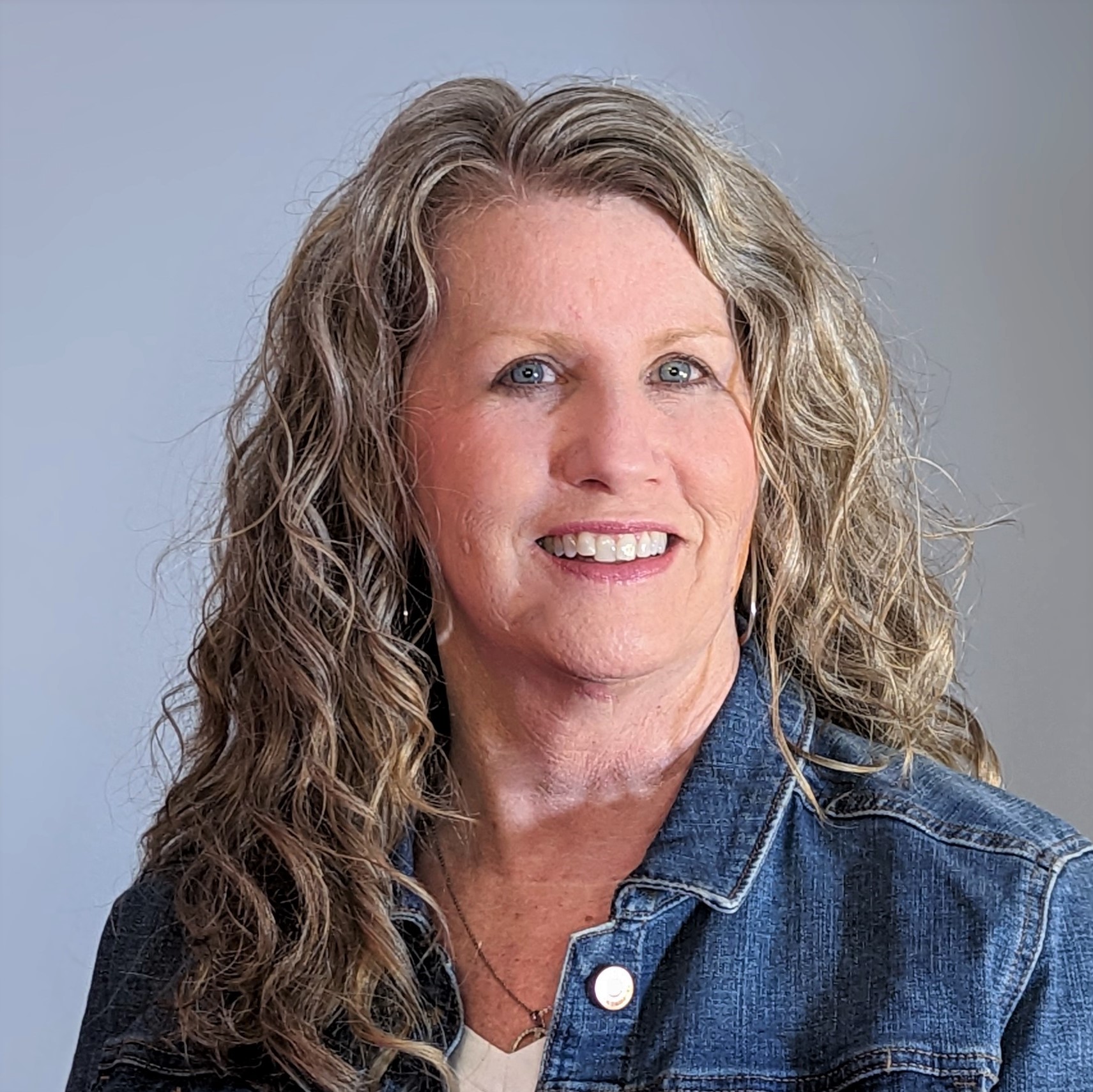 Tina Tharp is a White woman with long curly hair. She is wearing a denim jacket and smiling at the camera.