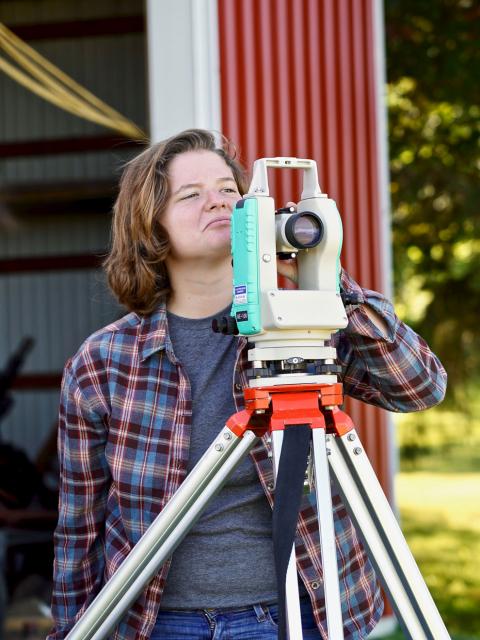 Esther Ramsey posing with surveying equipment near a barn